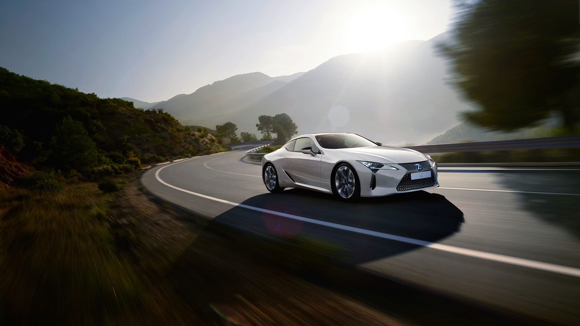 Hvit Lexus LC 500h sportssedan selvladende hybrid kommer kjørende oppover i et landskap med trær og fjell